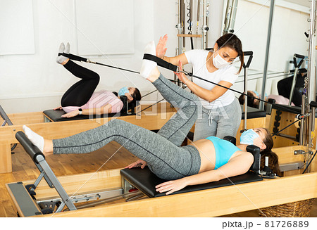 Female pilates trainer in protective mask helping pilates woman during training Female pilates trainer in protective mask helping pilates woman during training 81726889