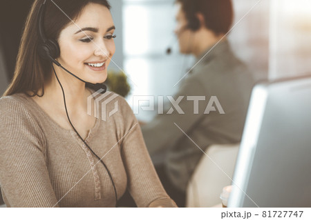 Young friendly girl in headsets is talking to a firm's client, while sitting at the desk in sunny office. Call center operators at work Young friendly girl in headsets is talking to a firm's client, while sitting at the desk in sunny office. Call center operators at work 81727747