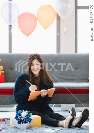 Asian young happy woman in furry jacket sitting in front gray couch with teddy bear dolls in living room smiling use pencil writing name list on orange notebook while wrapping present gift boxes Asian young happy woman in furry jacket sitting in front gray couch with teddy bear dolls in living room smiling use pencil writing name list on orange notebook while wrapping present gift boxes 81747470