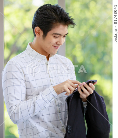 Asian successful professional happy handsome male employee officer standing smiling and using mobile smartphone holding suit jacket in hands in office behind working desk with computer monitor 81748041