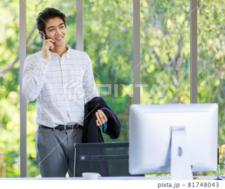 Asian successful professional happy handsome male employee officer standing smiling talking on mobile smartphone holding suit jacket in hands in office behind working desk with computer monitor 81748043