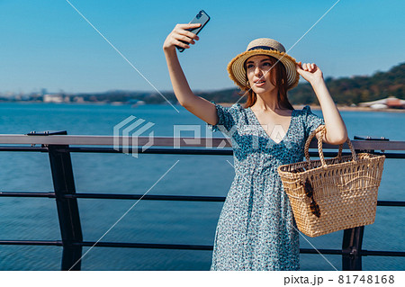 Attractive redhead woman posing at sunny day. Making selfie using phone. walks along the pier near the sea. Girl wearing fashionable blue dress and straw hat. Holding straw handbag 81748168