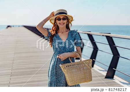 Beautiful redhead woman posing at sunny day. walks along the pier near the sea. Girl wearing fashionable sunglasses blue dress and straw hat. Holding straw handbag. smiling  81748217