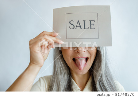 Playful lady with silver hair holds card with word Sale and shows tongue in studio Playful lady with silver hair holds card with word Sale and shows tongue in studio 81748570