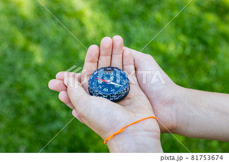 Girl's hands holding the compass. Hands of a teenager girl holding a liquid compass. Red compass needle points north. Green grass background. Copy space. Orienteering on the ground. 81753674