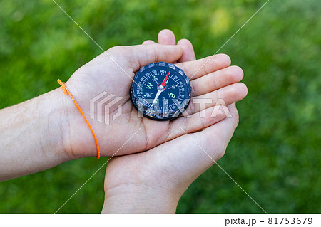 Girl holding the compass. Hands of a teenager girl holding a liquid compass. Green grass background. Copy space. Red compass needle points north. Orienteering on the ground. Selecting the direction 81753679