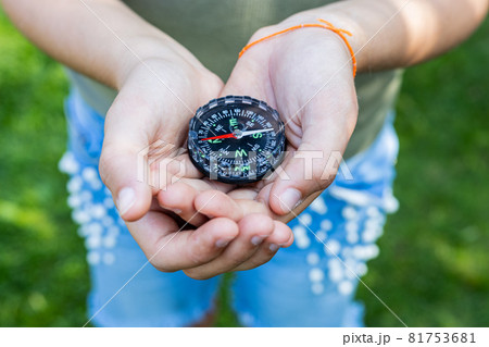 Girl holding the compass. Hands of a teenager holding a liquid compass. Red compass needle points north. Green grass background. Copy space. Orienteering on the ground. Selecting the direction 81753681