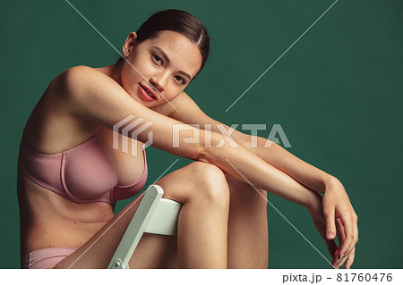 Cropped portrait of young caucasian woman in pink inner wear sitting on chair isolated over green studio background 81760476