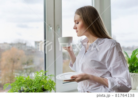 Portrait of young woman with cup of coffee near winter autumn window Portrait of young woman with cup of coffee near winter autumn window 81764138