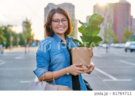 Middle aged woman with paper shopping bags with buying plant, looking at camera 81764711