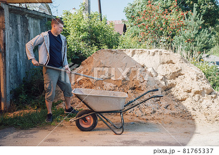 A 30-year-old man pours sand from a large pile of sand onto a garden wheelbarrow 81765337