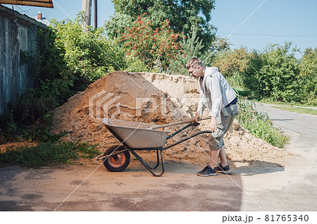 A 30-year-old man pours sand from a large pile of sand onto a garden wheelbarrow 81765340