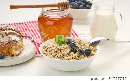 Morning breakfast, raw oatmeal flakes in a ceramic plate, milk in a decanter, blueberries and honey in a jar on a white table Morning breakfast, raw oatmeal flakes in a ceramic plate, milk in a decanter, blueberries and honey in a jar on a white table 81767755