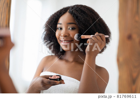 Natural makeup concept. Charming black lady applying blusher or powder in front of mirror at home 81769611