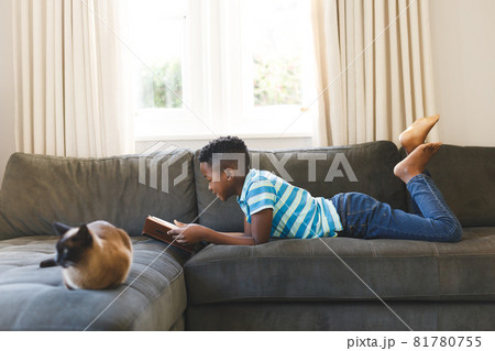 Happy african american boy reading book and lying on couch with cat in living room 81780755