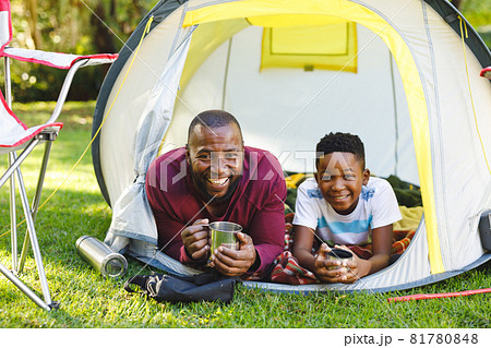 Portrait of african american father with son having fun and lying in tent with drinks in garden 81780848