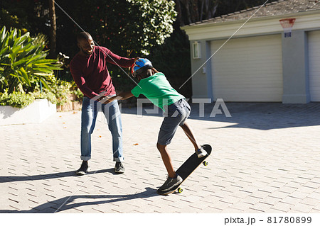African american father smiling and helping son balancing on skateboard in garden African american father smiling and helping son balancing on skateboard in garden 81780899