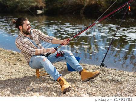 Positive man sitting near river and holding fishing rod 81791782