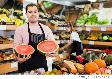 Young man seller holding half of watermelon in hands in fruit store Young man seller holding half of watermelon in hands in fruit store 81792174
