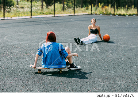 A young girl is sitting on a skateboard outdoors on a basketball court with her basketball player friend A young girl is sitting on a skateboard outdoors on a basketball court with her basketball player friend 81793376