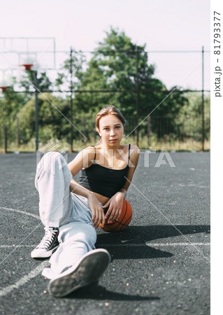 Portrait of a charming girl sitting on a sports field in a park or school with a basketball after a game or workout Portrait of a charming girl sitting on a sports field in a park or school with a basketball after a game or workout 81793377