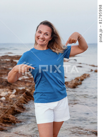 Middle aged woman pointing at t-shirt and standing on the cliff Middle aged woman pointing at t-shirt and standing on the cliff 81795998