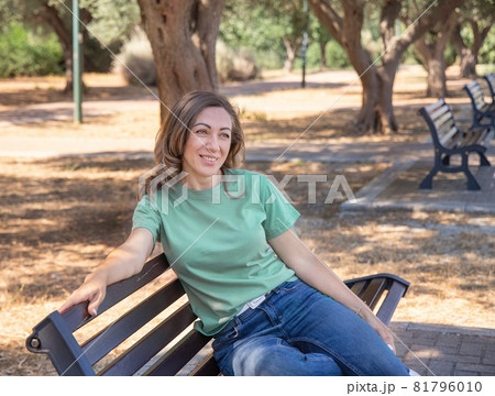 Women wearing t-shirt and jeans sits on the bench in the park Women wearing t-shirt and jeans sits on the bench in the park 81796010