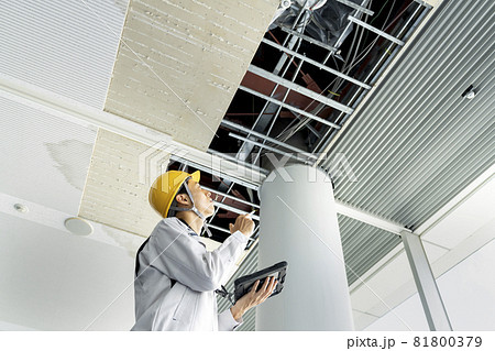 タブレットを持ってビル天井の破損箇所を調査する作業服姿の男性　 81800379