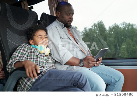 Afro American boy with headphones around neck sleeping on fathers shoulder in bus while he using tablet Afro American boy with headphones around neck sleeping on fathers shoulder in bus while he using tablet 81809490