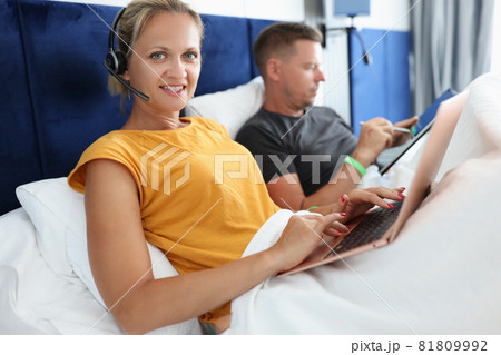Woman with headphones with microphone lying in bed with laptop next to her husband 81809992