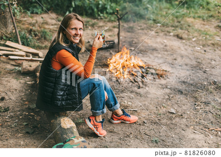A young woman is warming herself by the fire with a cup of warming tea in the forest 81826080
