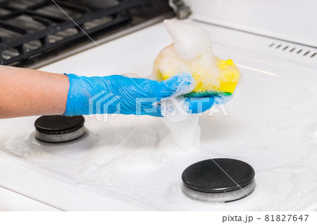 Woman's hand in a household glove washing sponge with foam a dirty gas stove background, close up 81827647