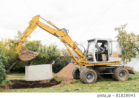 Hydraulic piston system of the excavator with a bucket, lowering into the pit on steel cables concrete sewer ring. Construction or repair of sewer home Hydraulic piston system of the excavator with a bucket, lowering into the pit on steel cables concrete sewer ring. Construction or repair of sewer home 81828043