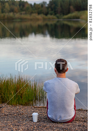A young man is sitting on the shore of the lake and looking into the distance, next to a white mug of coffee or tea. Solitude and relaxation in nature. A beautiful landscape of a lake in the forest  81832798