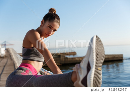 Close-up of attractive sportswoman holding leg on handrope, stretching on the seaside promenade. Female runner warming-up for morning jogging training 81848247