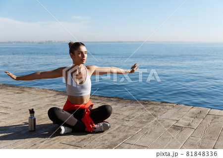 Outdoor shot of relaxed young woman sitting on wooden pier and looking at the sea, doing yoga exercises 81848336