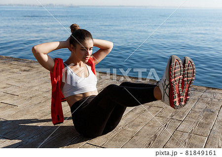 Outdoor shot of confident fitness woman working on her abs. Sportswoman doing crunches with raised legs, training near sea on a pier Outdoor shot of confident fitness woman working on her abs. Sportswoman doing crunches with raised legs, training near sea on a pier 81849161