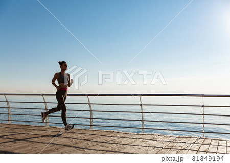 Side view of young fitness woman running along pier. Female athlete workout and jogging on the seaside promenade 81849194