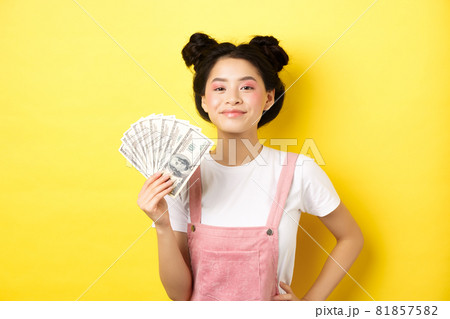 Shopping. Smiling asian girl with stylish makeup, showing dollar bills with pleased face, making money, standing on yellow background 81857582