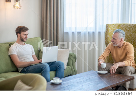Young bearded man working on a laptop, his father drinking tea Young bearded man working on a laptop, his father drinking tea 81863364