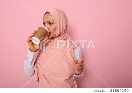 Beautiful Arabic muslim woman with covered head drinking hot drink, tea or coffee from disposable cardboard takeaway cup, standing sideways, three quarters, against pink background with copy space Beautiful Arabic muslim woman with covered head drinking hot drink, tea or coffee from disposable cardboard takeaway cup, standing sideways, three quarters, against pink background with copy space 81864190