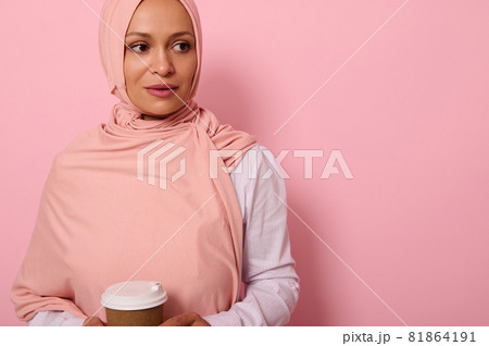 Close-up. Arab Muslim beautiful woman wearing pink hijab and white shirt holding recyclable disposable ecological paper mug in her hands, looking the side posing on colored background with copy space 81864191