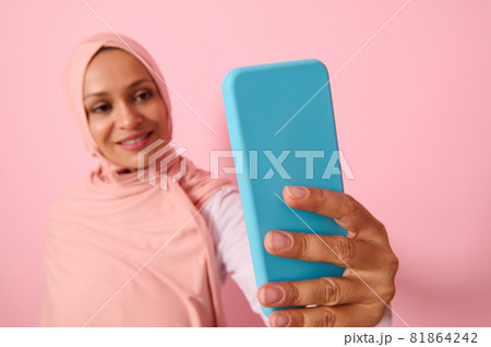 Soft focus on the smartphone in blue cover in outstretched arms of Arab Muslim woman wearing traditional religious islamic outfit, pink hijab,and smiles with toothy smile making self-portrait, selfie Soft focus on the smartphone in blue cover in outstretched arms of Arab Muslim woman wearing traditional religious islamic outfit, pink hijab,and smiles with toothy smile making self-portrait, selfie 81864242