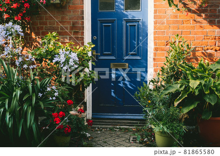 Blue front door and brick walls facade of a Beautiful Cottage with many various potted flowers. The Small garden in front of the house, gardening hobby, create a cozy space around Blue front door and brick walls facade of a Beautiful Cottage with many various potted flowers. The Small garden in front of the house, gardening hobby, create a cozy space around 81865580