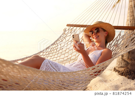 Working at sunset. Young happy female using smartphone while lying in the hammock on the beach 81874254