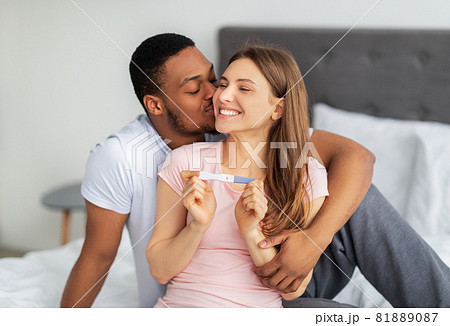 Happy multinational couple celebrating positive result of pregnancy test on bed at home Happy multinational couple celebrating positive result of pregnancy test on bed at home 81889087