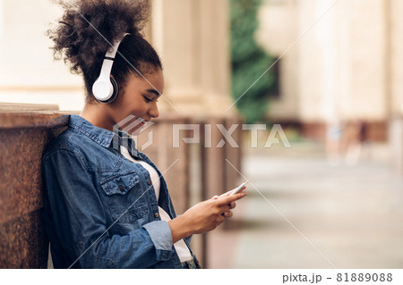 African Female Girl Using Smartphone Wearing Earphones At College Outdoors African Female Girl Using Smartphone Wearing Earphones At College Outdoors 81889088
