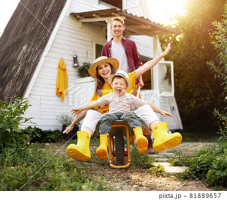 cheerful family of farmers enjoying holiday and having fun. mother and father roll their kid boy in a garden wheelbarrow in the summer cheerful family of farmers enjoying holiday and having fun. mother and father roll their kid boy in a garden wheelbarrow in the summer 81889657
