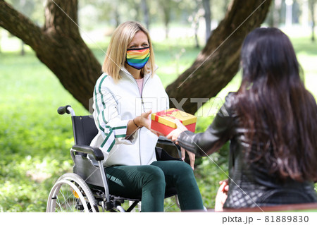 Woman giving gift in red box to disabled friend in protective medical mask with lgbt symbols in park 81889830