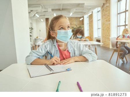 Gorgeous little girl wearing protective face mask during coronavirus outbreak looking away and listening attentively. Kid studying in elementary school, sitting at the desk Gorgeous little girl wearing protective face mask during coronavirus outbreak looking away and listening attentively. Kid studying in elementary school, sitting at the desk 81891273